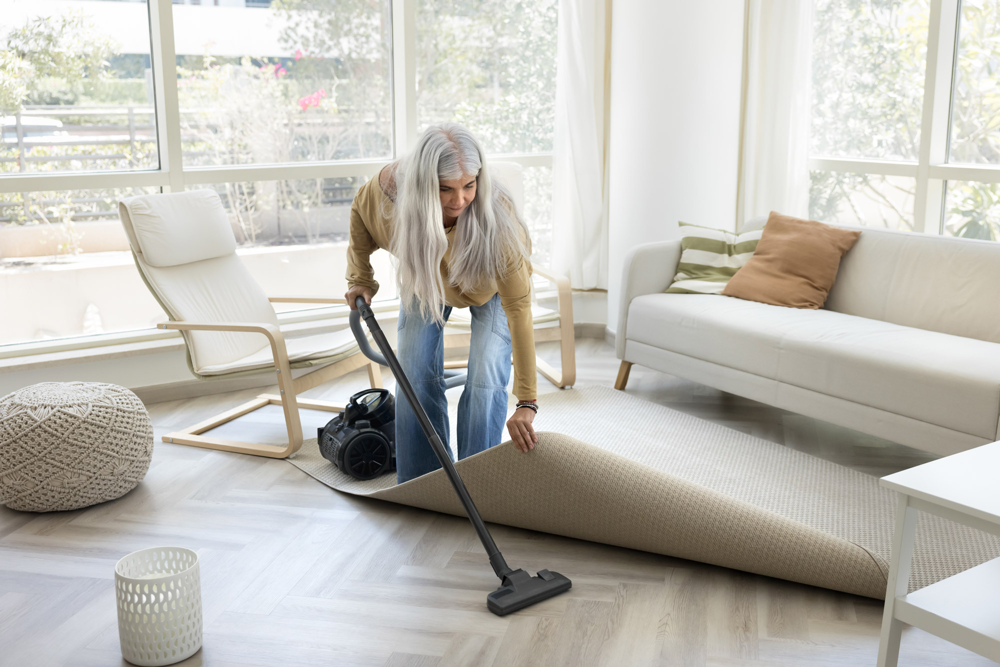 Woman with long grey hair vacuums under a rug in a bright, modern living room